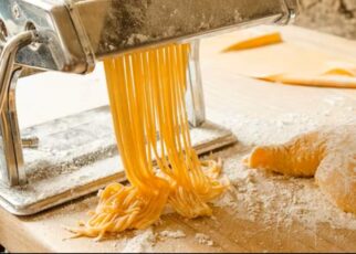 golden fresh pasta strands emerging from an electric pasta maker, dusted with flour on a wooden kitchen counter