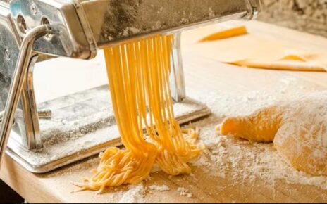 golden fresh pasta strands emerging from an electric pasta maker, dusted with flour on a wooden kitchen counter