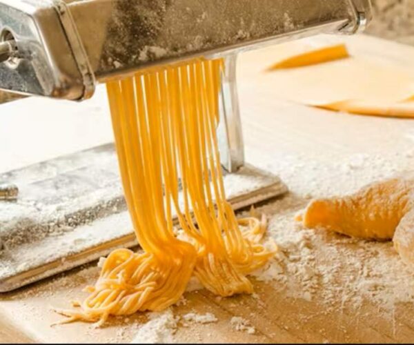 golden fresh pasta strands emerging from an electric pasta maker, dusted with flour on a wooden kitchen counter